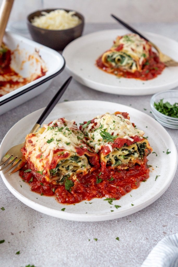 two spinach lasagna rolls on a white plate with marinara sauce and another plate with lasagna in the background