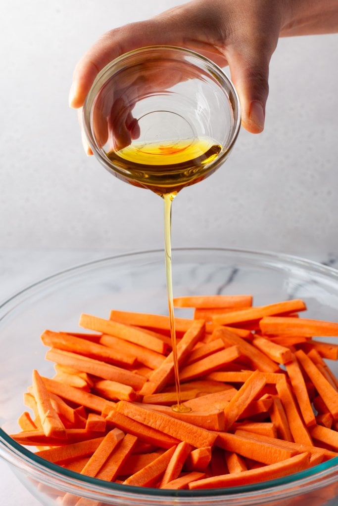 pouring olive oil over sweet potatoes in a glass bowl