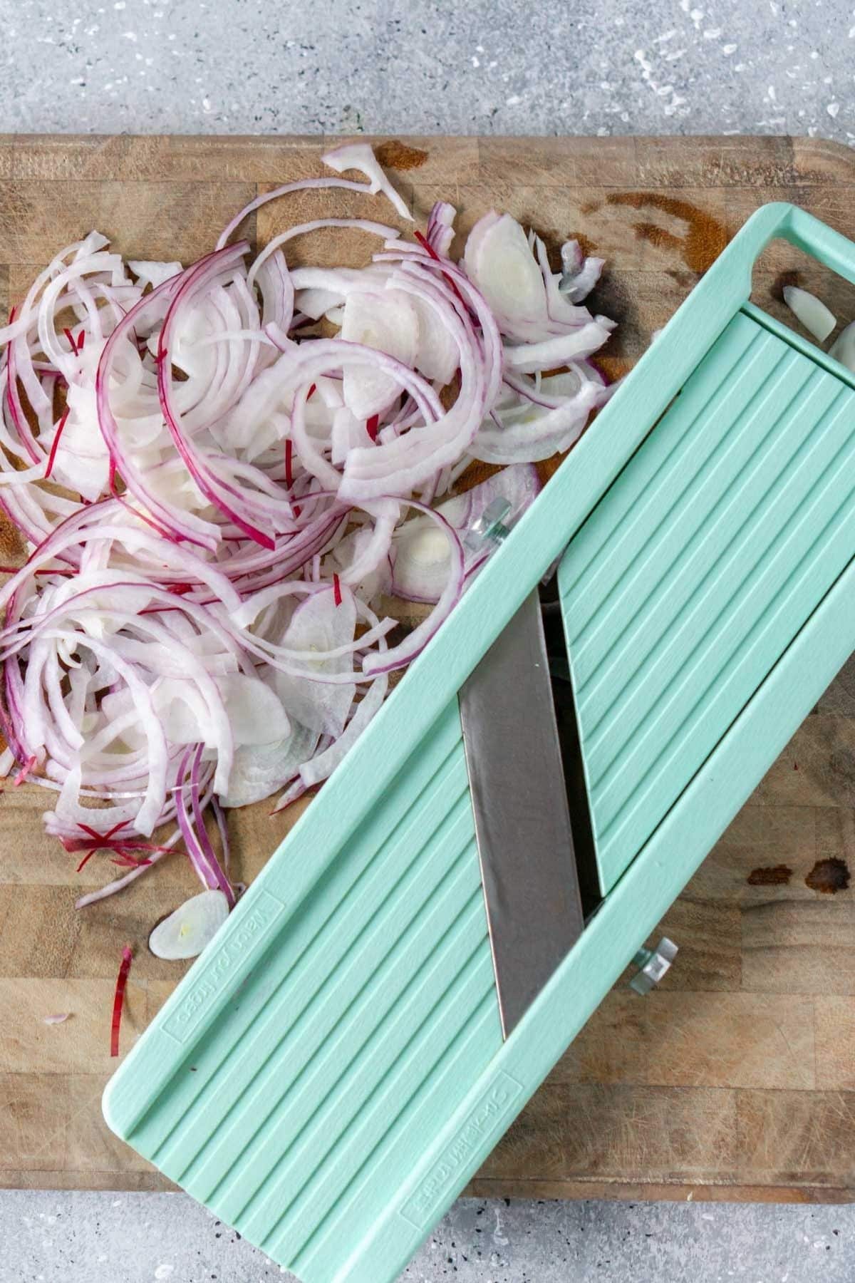 Red onion slices lay on a countertop next to a mandoline.