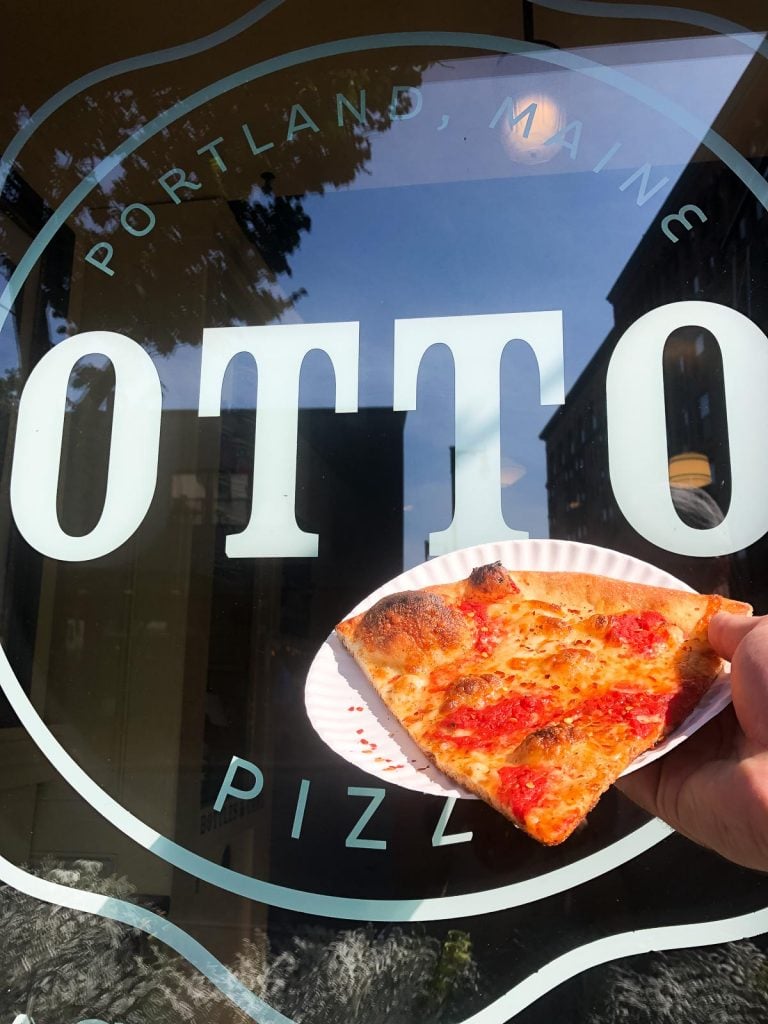 a hand holding a slice of pizza in front of the otto pizza sign