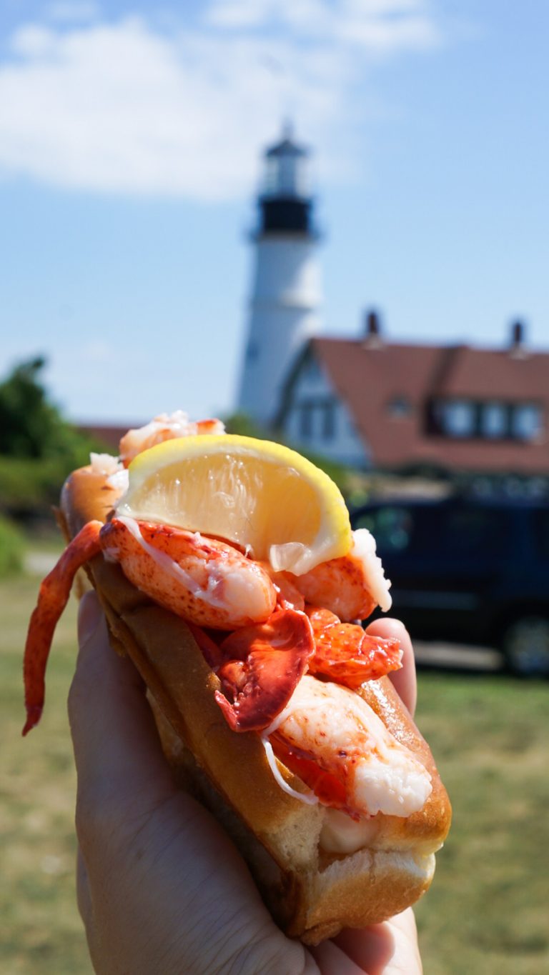 a hand holding a lobster roll with a lighthouse in the background