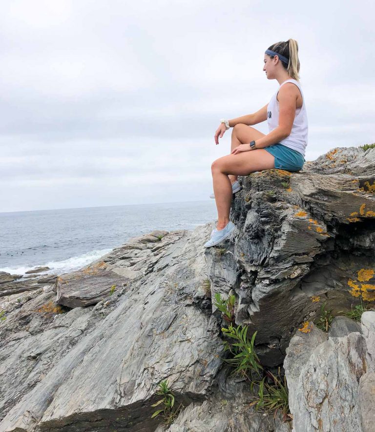 a girl with blonde hair sitting on the edge of a rock in shorts and a white shirt