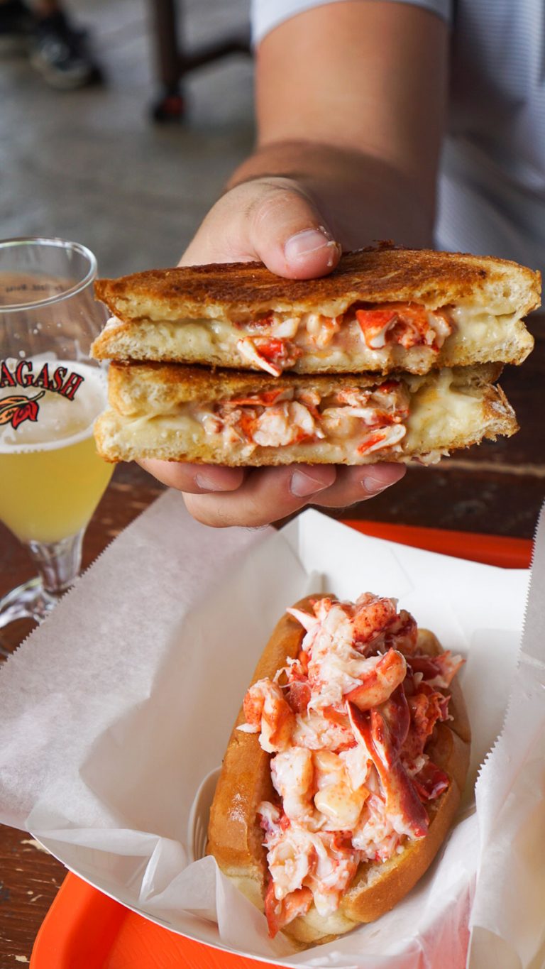 a hand holding a lobster grilled cheese with a beer and a lobster roll