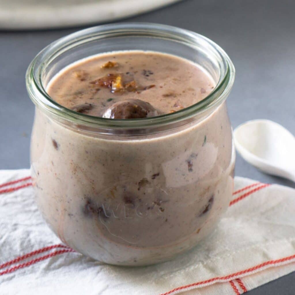 Bacon mushroom gravy in a clear glass jar atop a striped dish towel with a white spoon on the side.