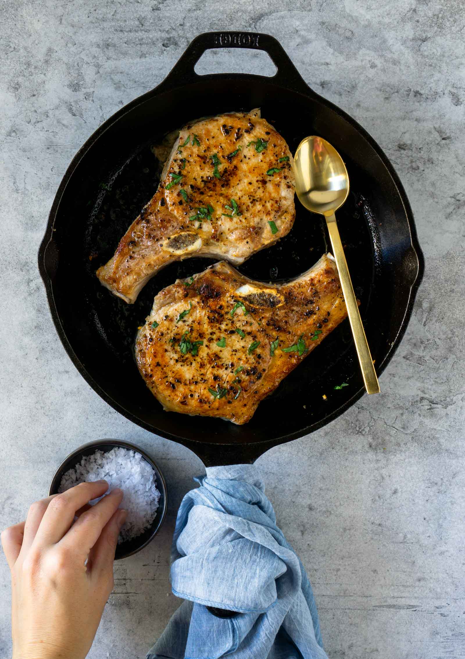 two pork chops in a black cast iron pan with a gold spoon and a hand reaching for salt