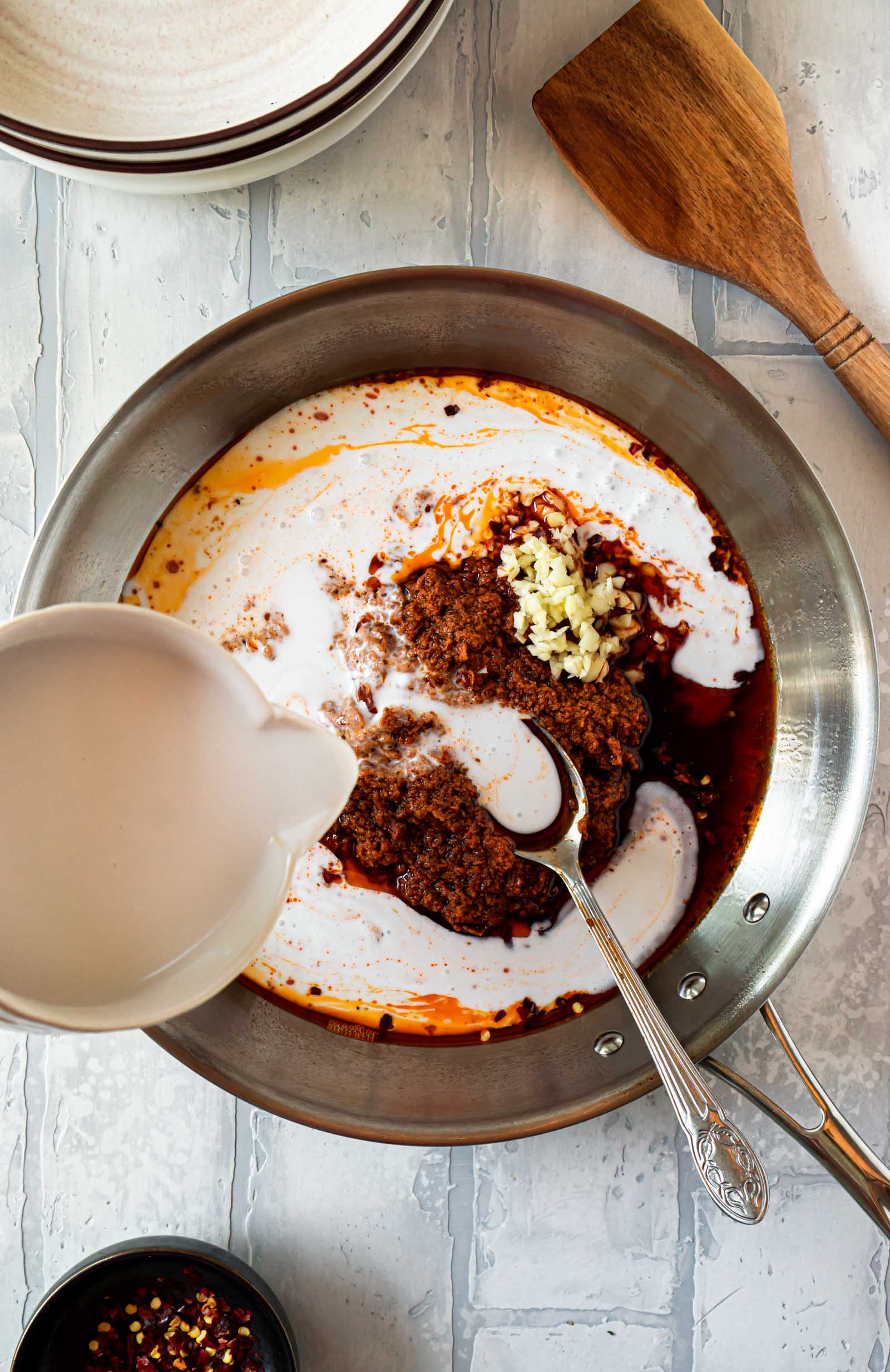 coconut cream being poured into a pan with sun dried tomato pesto and garlic, and a silver spoon