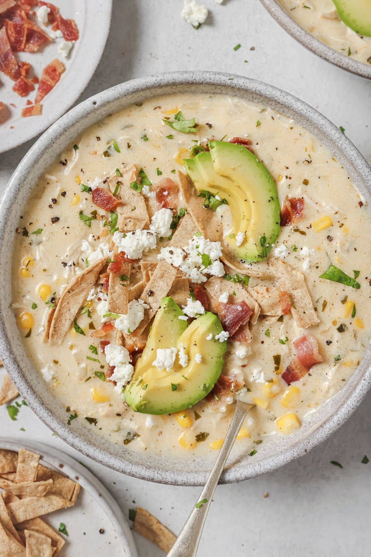 A bowl full of white chicken chili topped with bacon, cheese and avocado slices.