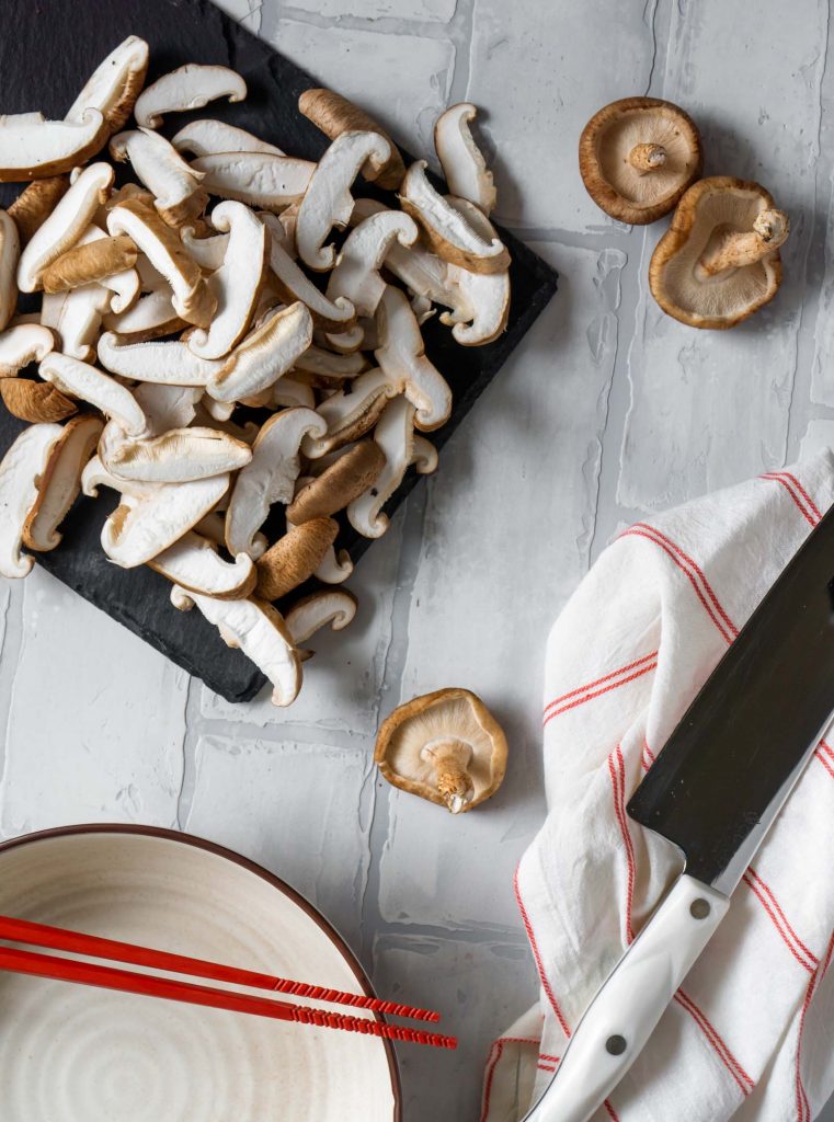 sliced shiitake mushrooms on a black slate board, red chopsticks, and a knife