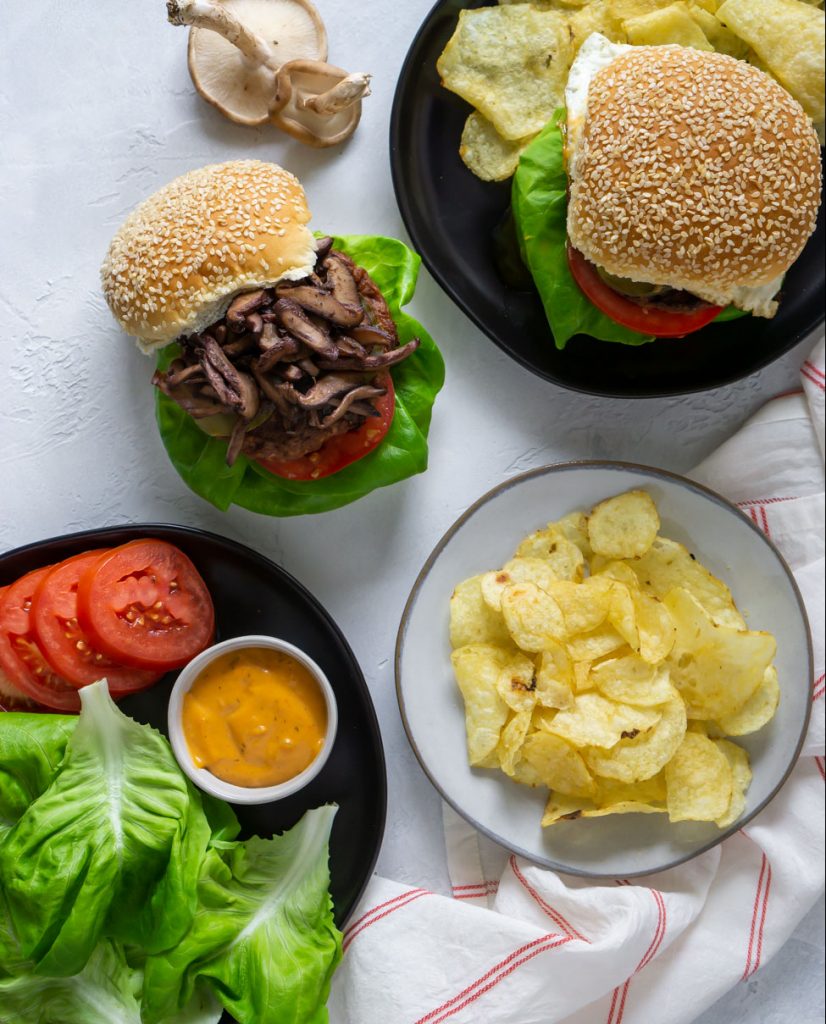 two burgers, chips on a white plate, lettuce and tomato on a black plate