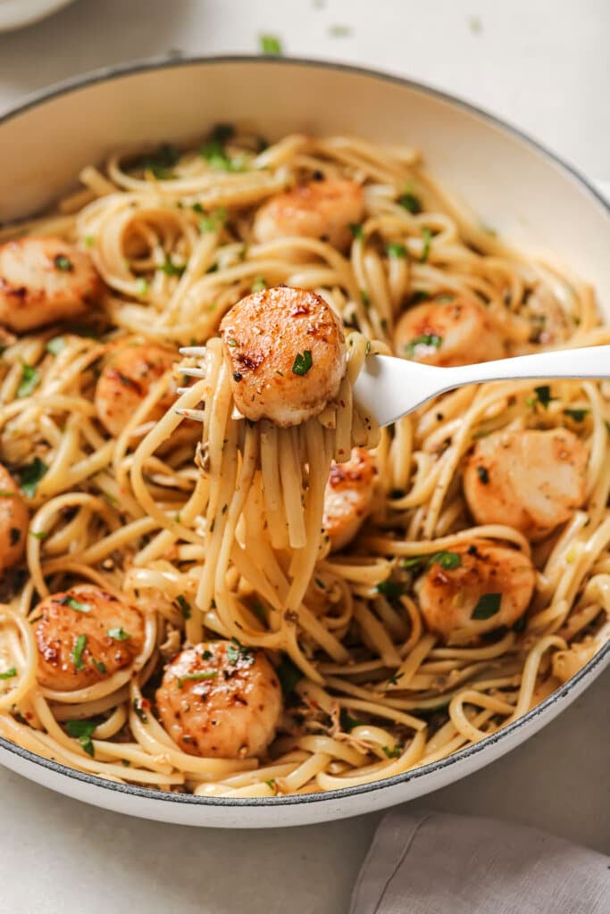 a white fork lifting a bite of linguine with clam sauce and a seared scallop on top