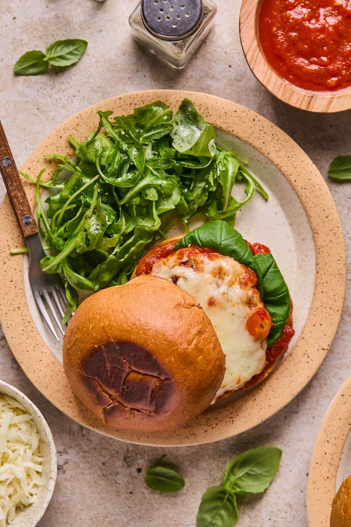 Chicken parmesan burger with a toasted bun alongside a salad, a bowl of sauce, and shredded cheese.