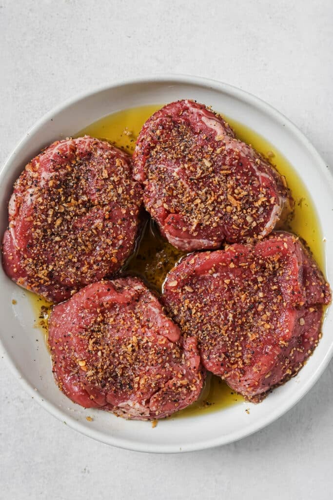 Four seasoned filet mignon steaks in a white bowl, coated in oil and seasoning.