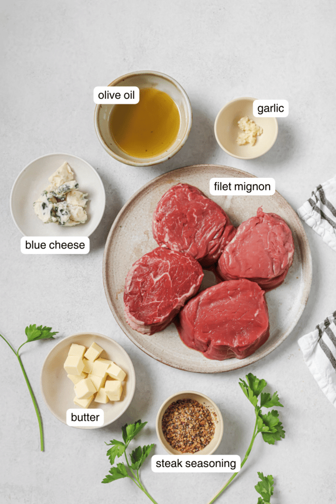 four steaks on a white plate, butter, blue cheese, steak seasoning, and olive oil in small ingredient bowls