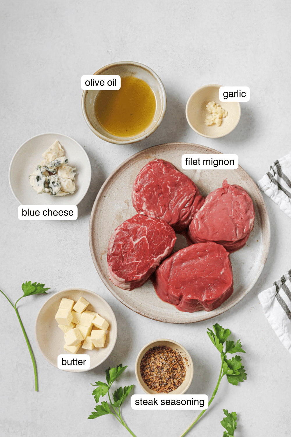 Four steaks on a white plate, butter, blue cheese, steak seasoning, and olive oil in small ingredient bowls.