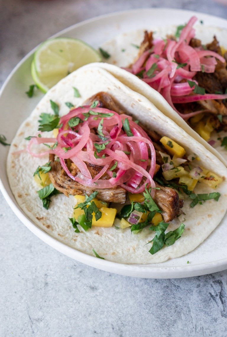 two carnitas tacos on a white plate with lime wedges