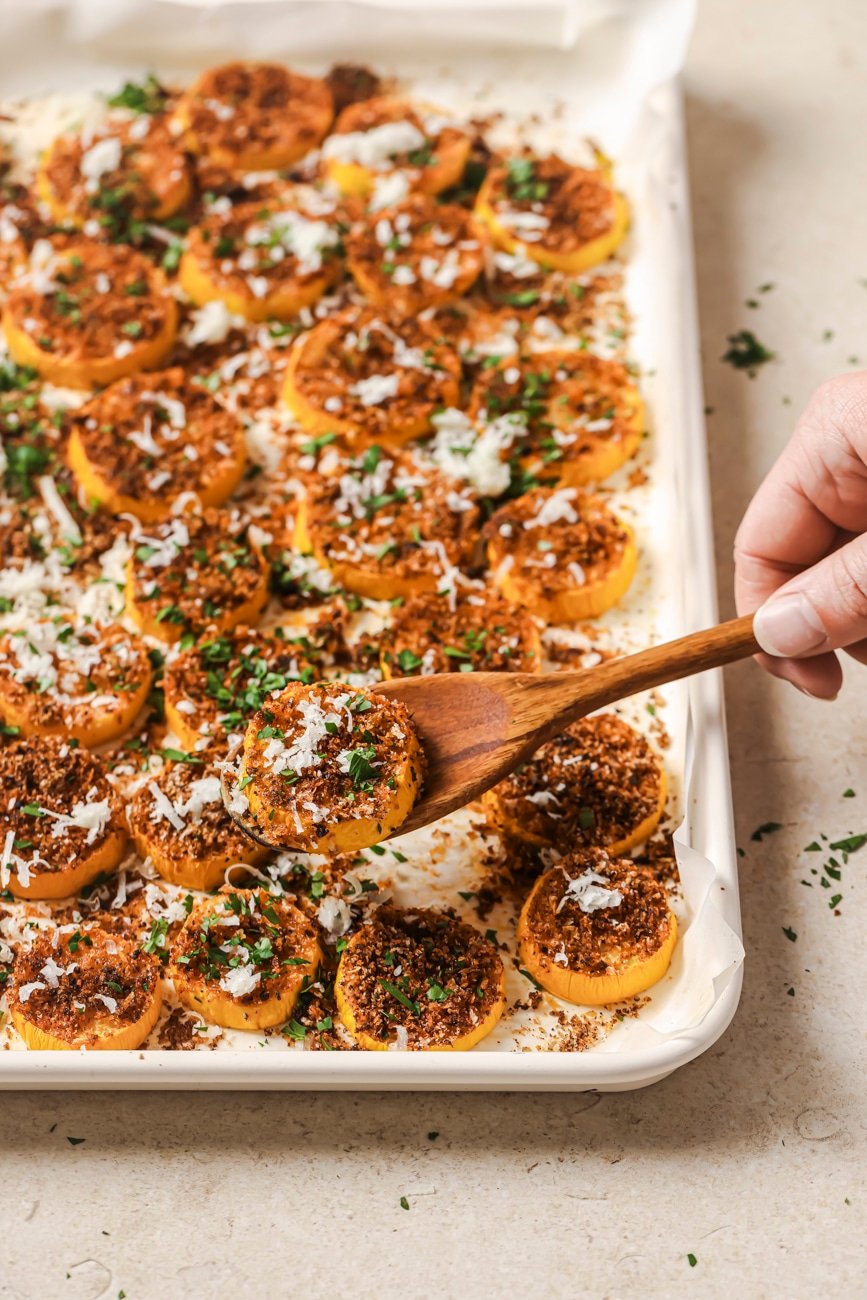 a wooden spoon holding a piece of roasted yellow squash over a sheet pan full of cooked squash.