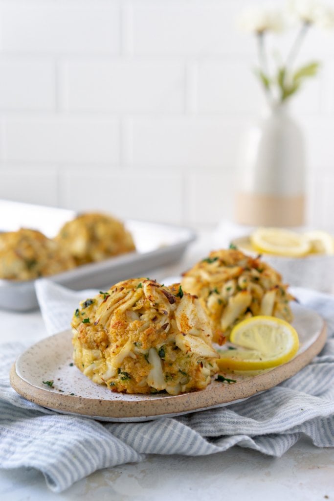 two maryland crab cakes on a plate with a sheet pan and flowers in the background