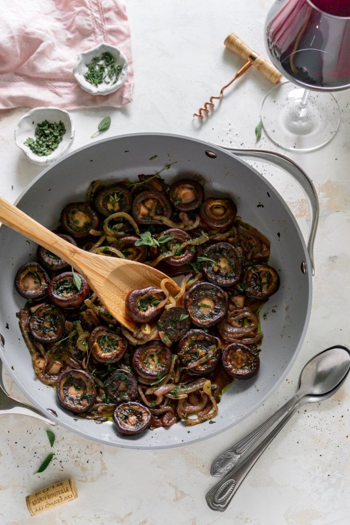 red wine mushrooms in a pan with a glass of wine, herbs, and spoons