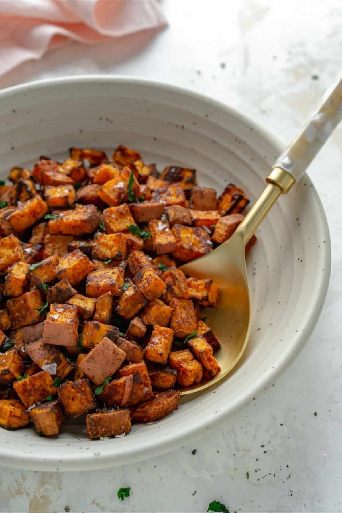 Cubed roasted sweet potatoes in a ceramic bowl with a gold spoon.