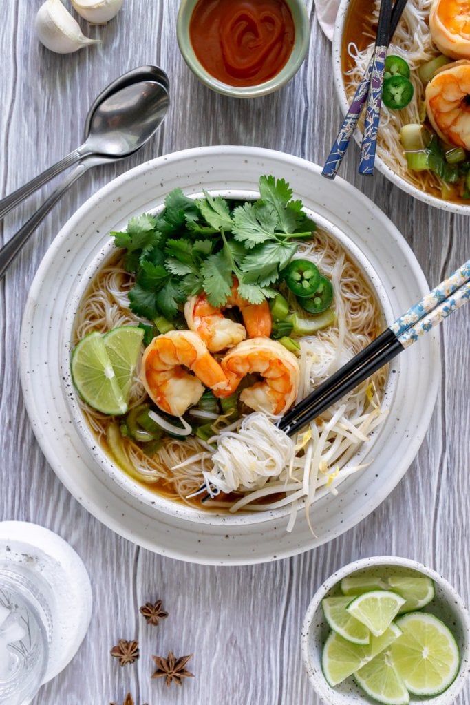 two bowls of shrimp pho noodle soup with chopsticks, sriracha, and limes
