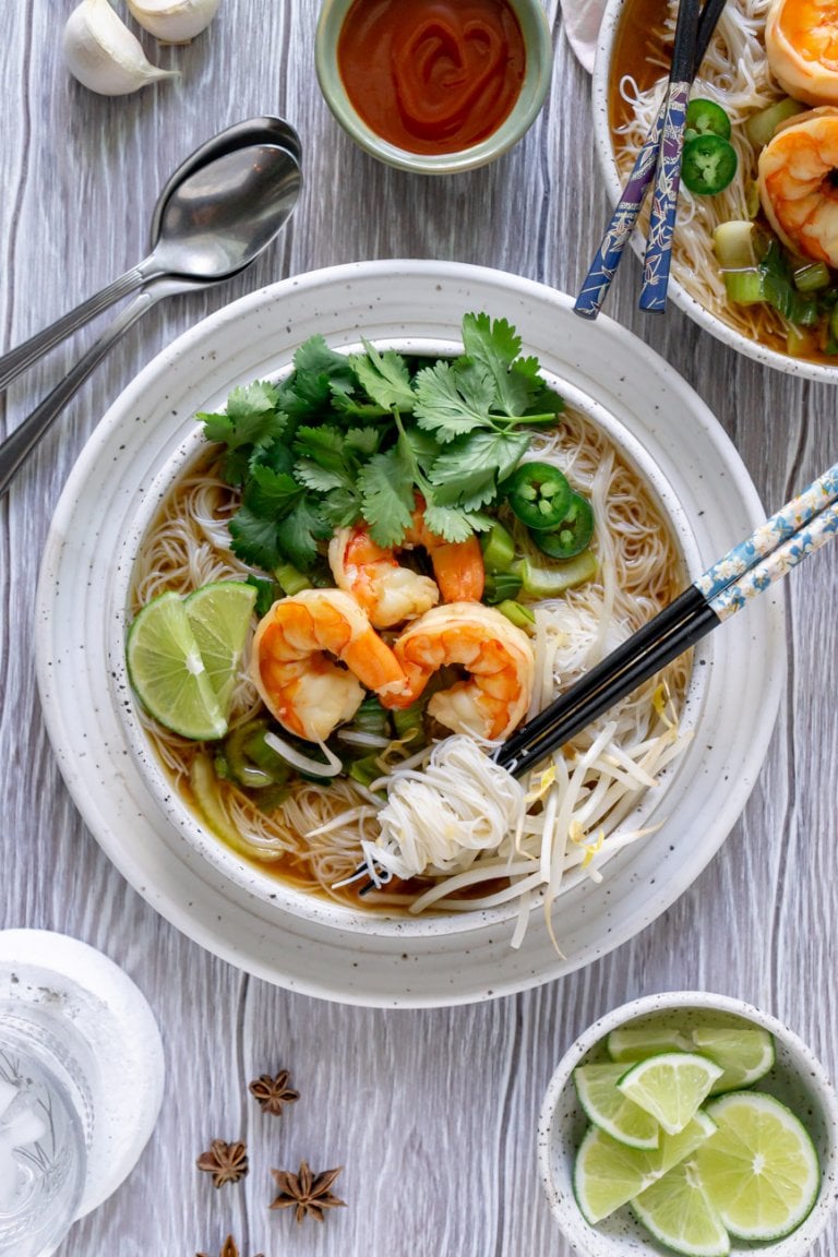 two bowls of shrimp pho noodle soup with chopsticks, sriracha, and limes