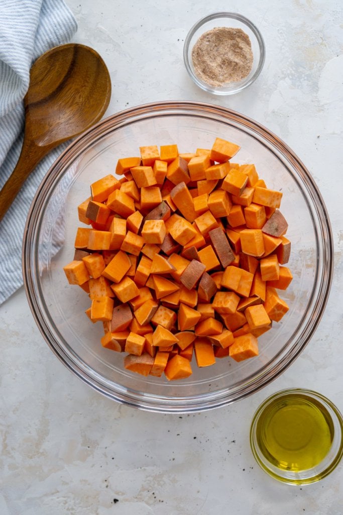 cubed sweet potatoes in a glass bowl with a wooden spoon, seasoning, and oil