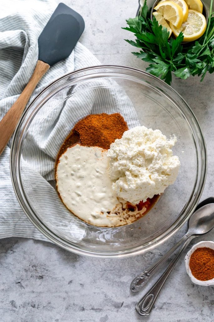 old bay, tartar sauce, and cream cheese in a clear glass mixing bowl with a blue linen, parsley, lemon, and spatula on the side.