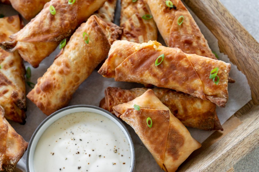 Cooked buffalo chicken egg rolls in a wooden serving tray and garnished with sliced green scallions, with a cup of ranch dressing.