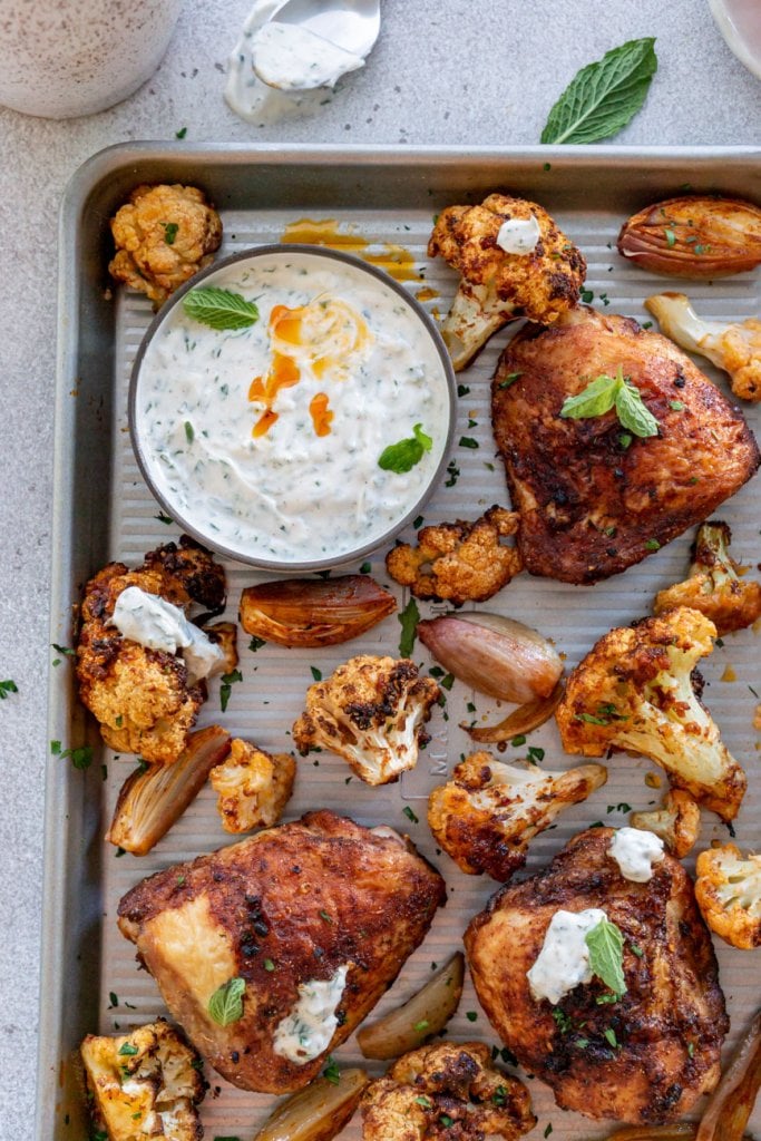 baked chicken and cauliflower on a pan with a bowl of yogurt sauce