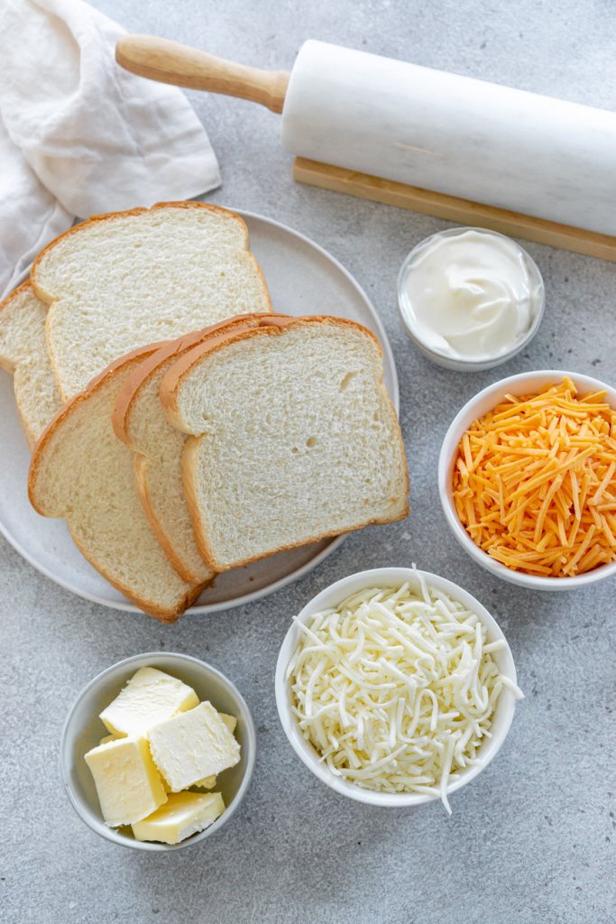 white bread on a white plate, shredded cheese in bowls, cubed butter in small bowl, and a rolling pin