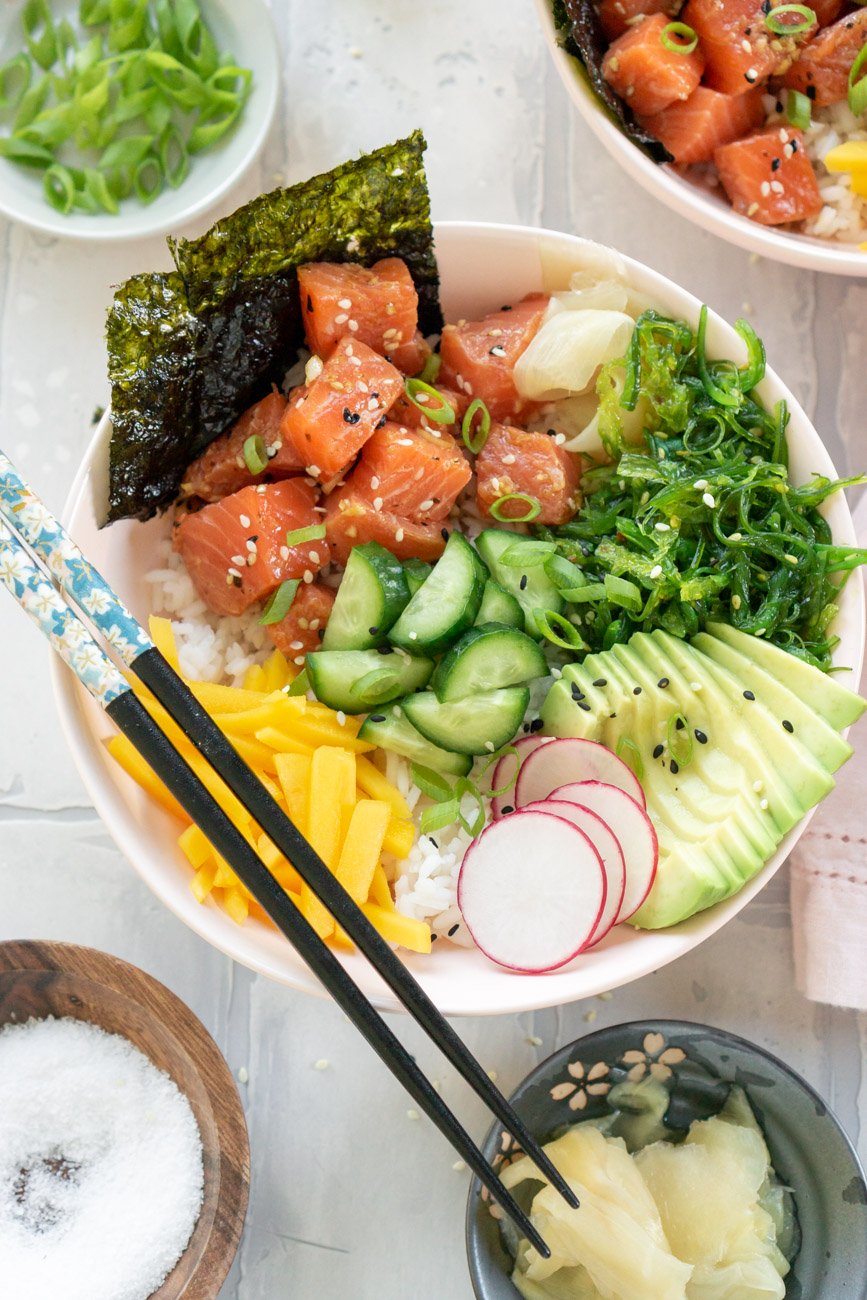 Salmon poke bowl with chopsticks, ginger, and scallions to the side.