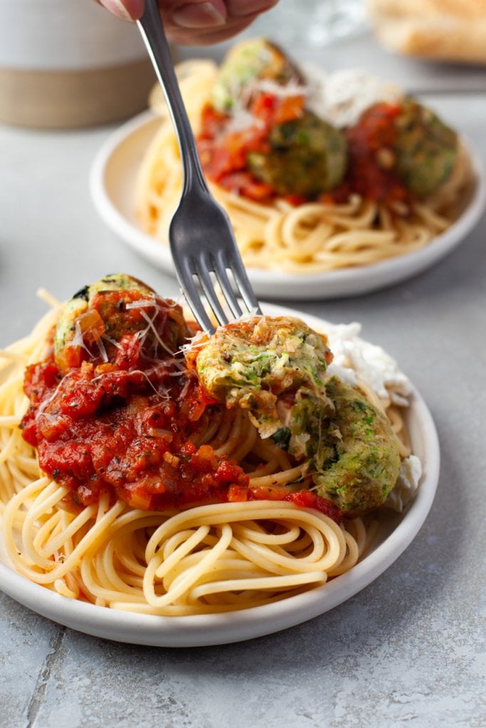 two plates of zucchini meatballs with spaghetti and a fork taking a bite