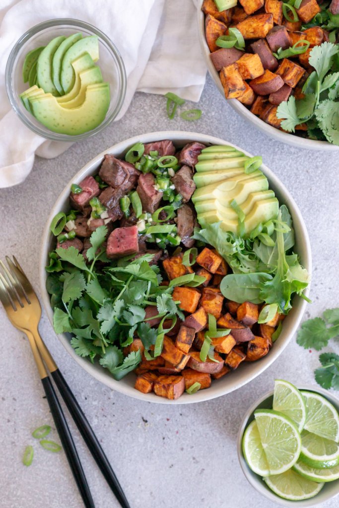 two steak bowls with forks, avocado, and sliced limes to the side