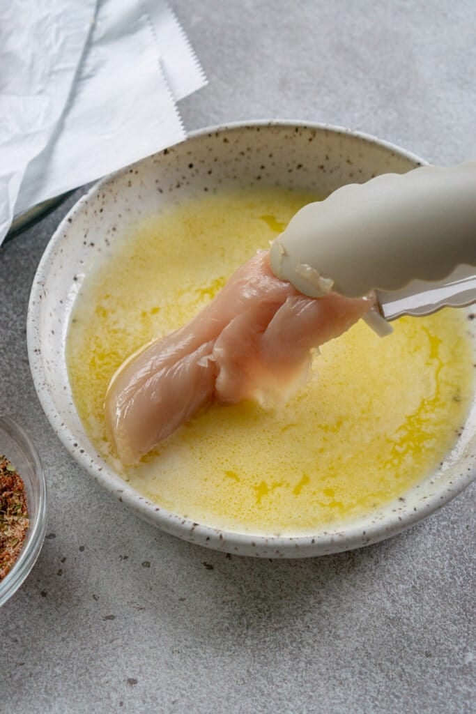 A chicken tender being dipped into a bowl of melted butter with tongs.