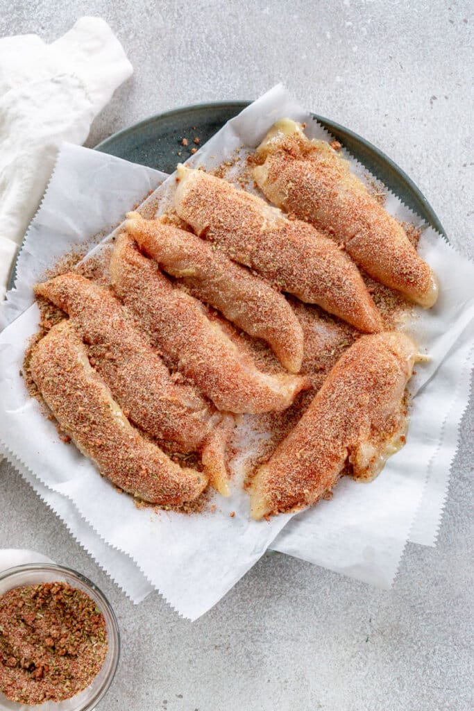 Seasoned chicken tenders on a plate lined with parchment paper.