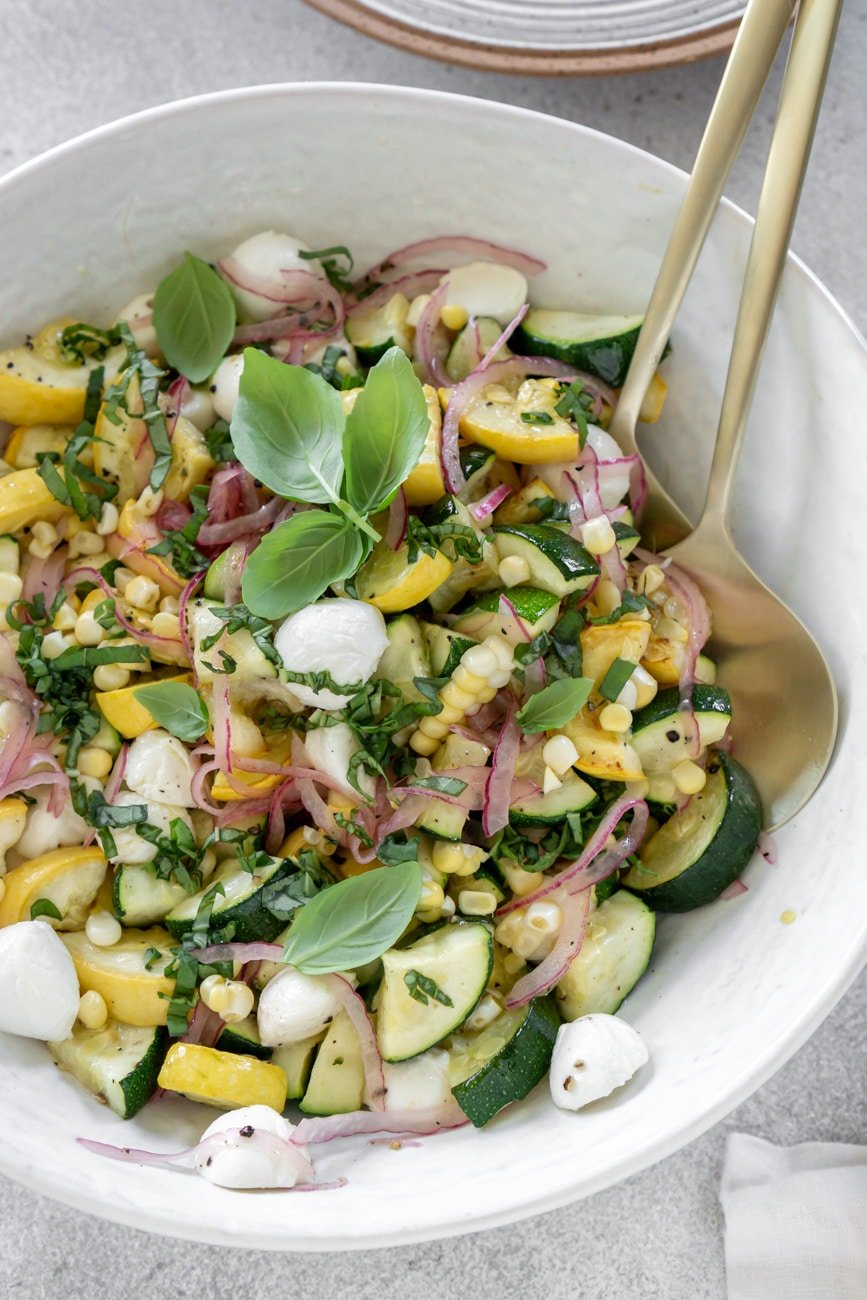 Zucchini salad in a large white bowl with gold serving spoons.