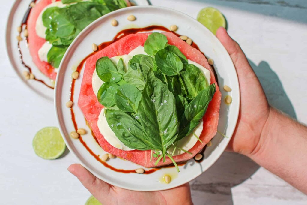 Watermelon mozzarella salad on a white plate held by two hands with basil on top.