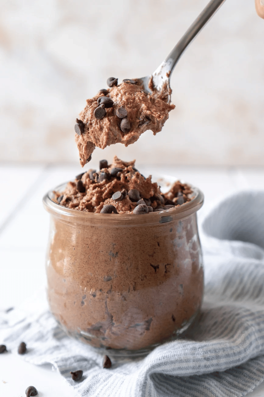 Spoon holding up a scoop of brownie batter cookie dough above clear jar.