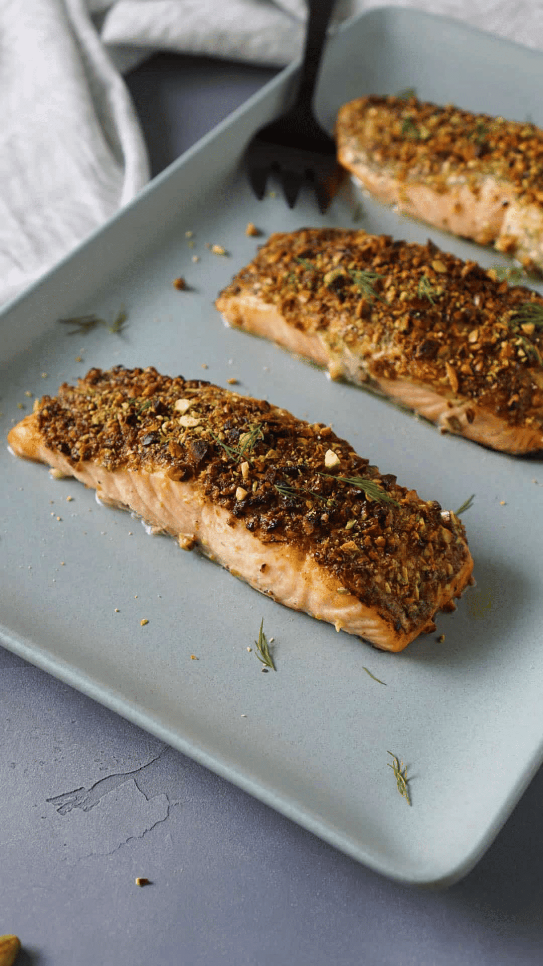 Three pieces of cooked pistachio crusted salon on a baking sheet with herbs.