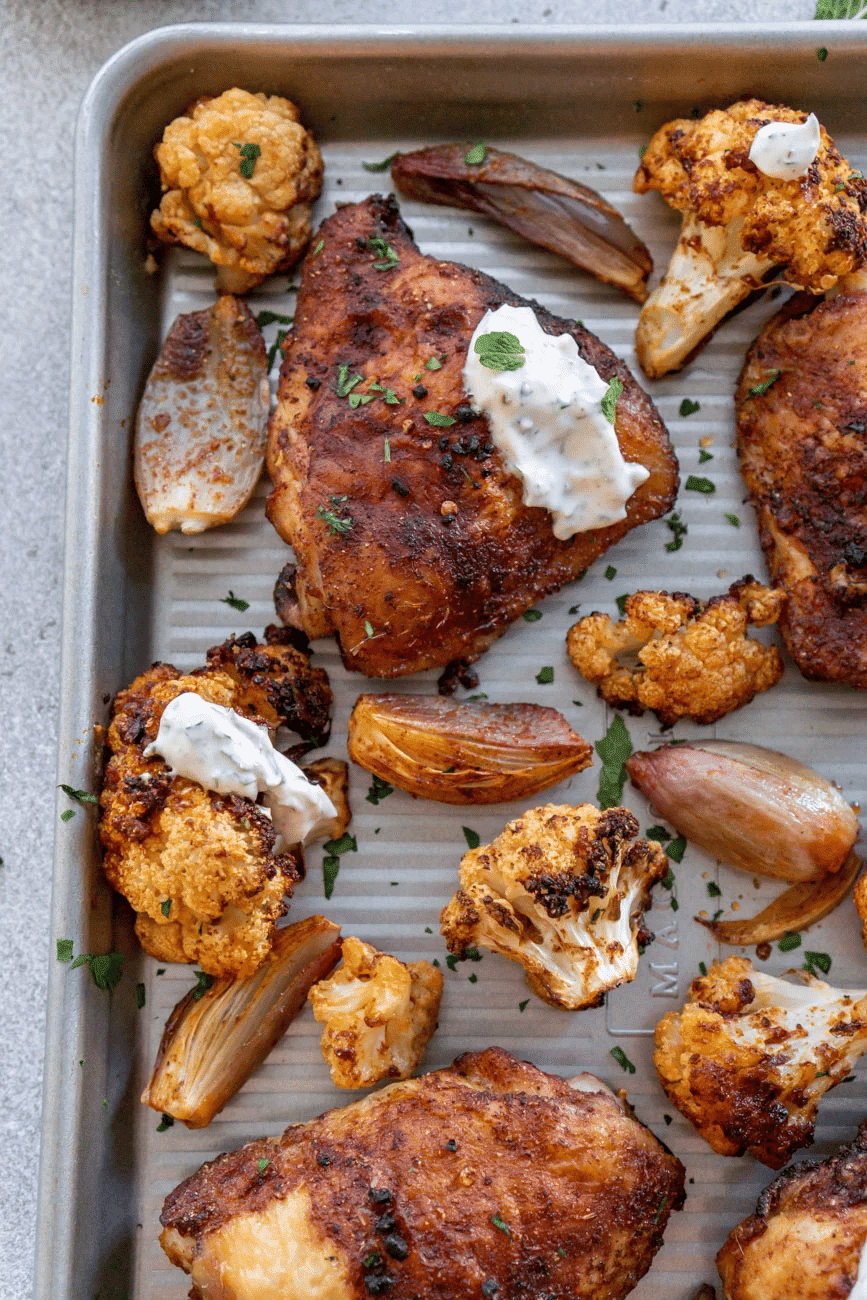 Chicken, onions and cauliflower on a baking sheet with herbed yogurt sauce on chicken.