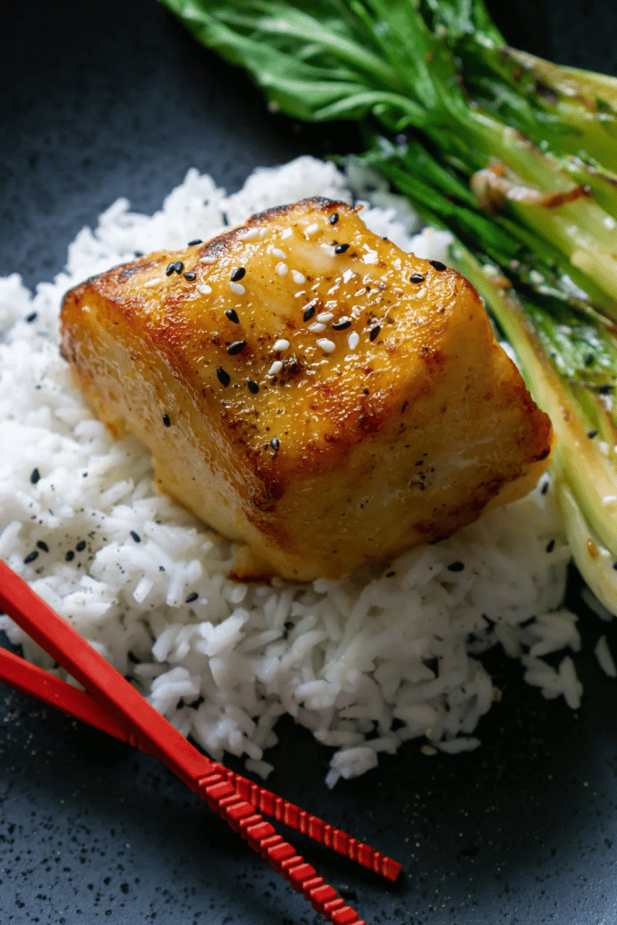 close up of miso glazed chilean sea bass on top of rice with vegetables in background 