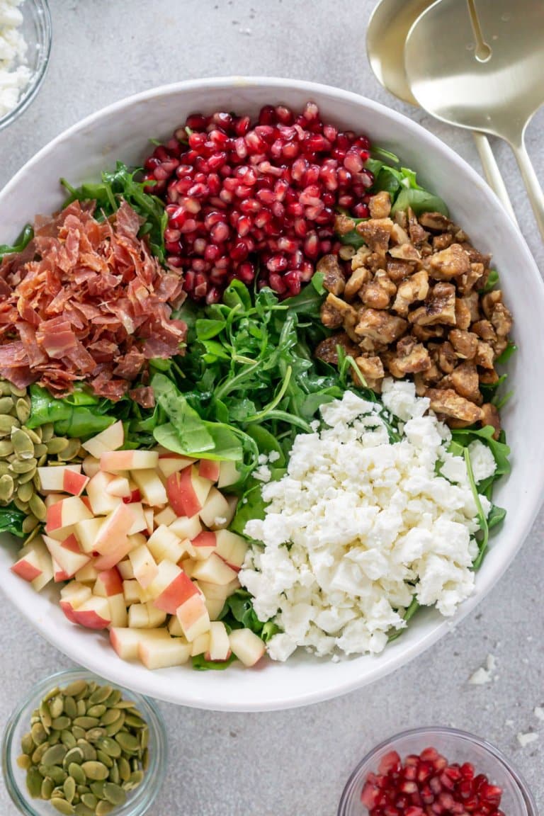 a bowl of lettuce with pomegranate, feta cheese, apples, prosciutto, and walnuts