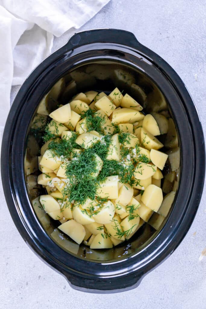 Uncooked chopped potatoes in a slower cooker, topped with dill and garlic.