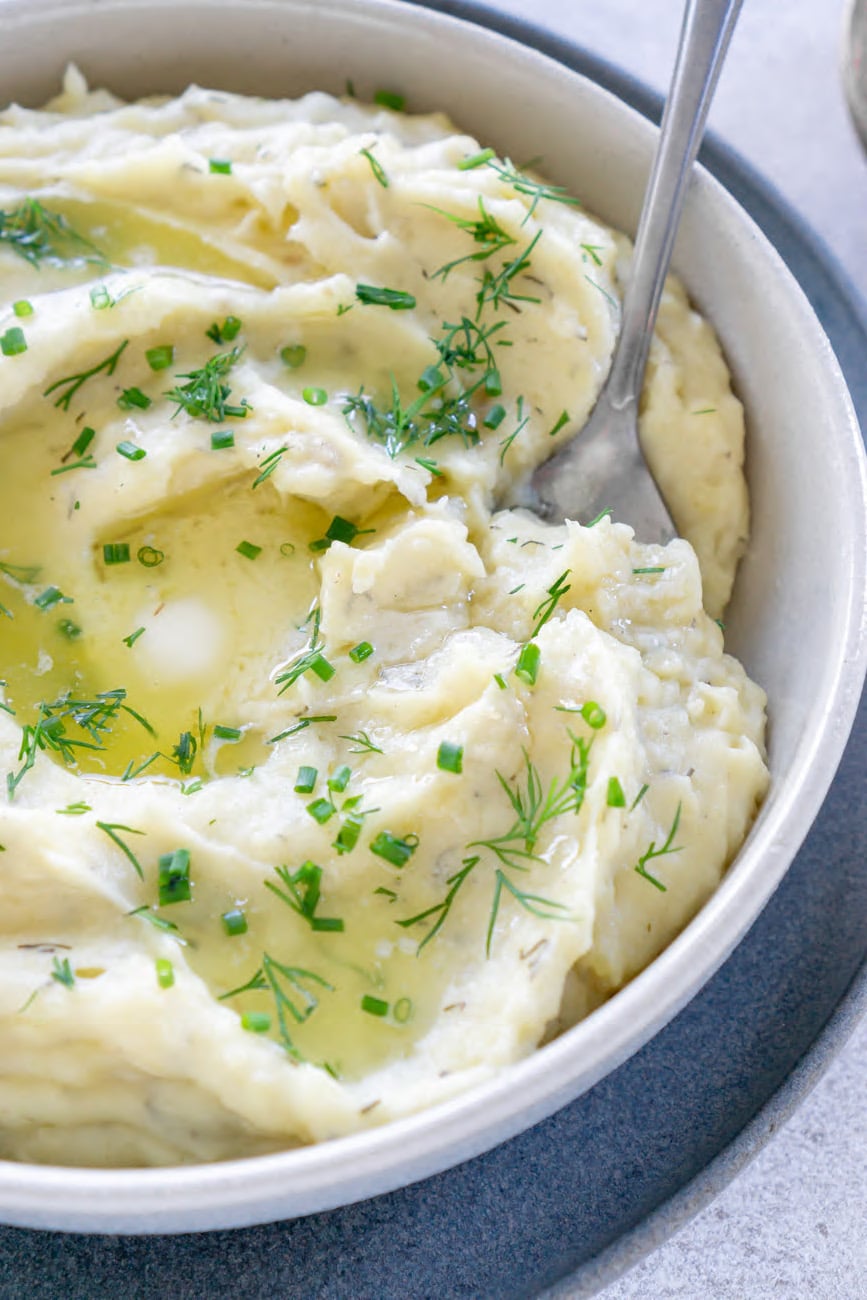 A bowl of mashed potatoes topped with chives and dill, with a spoon.