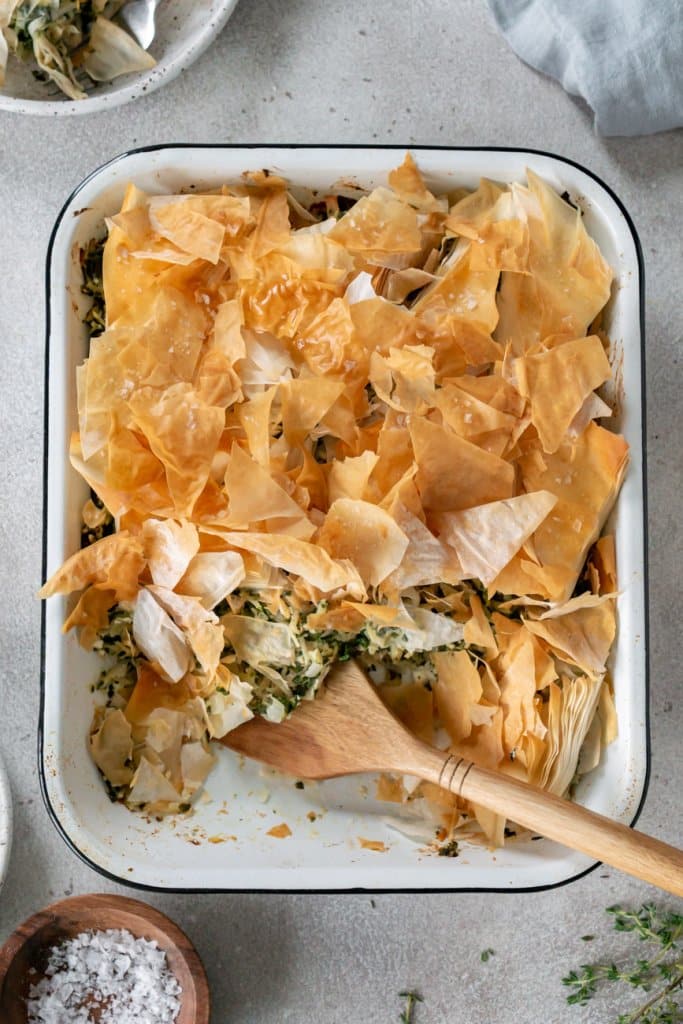 spanakopita chicken bake in a white baking dish and a wooden spatula
