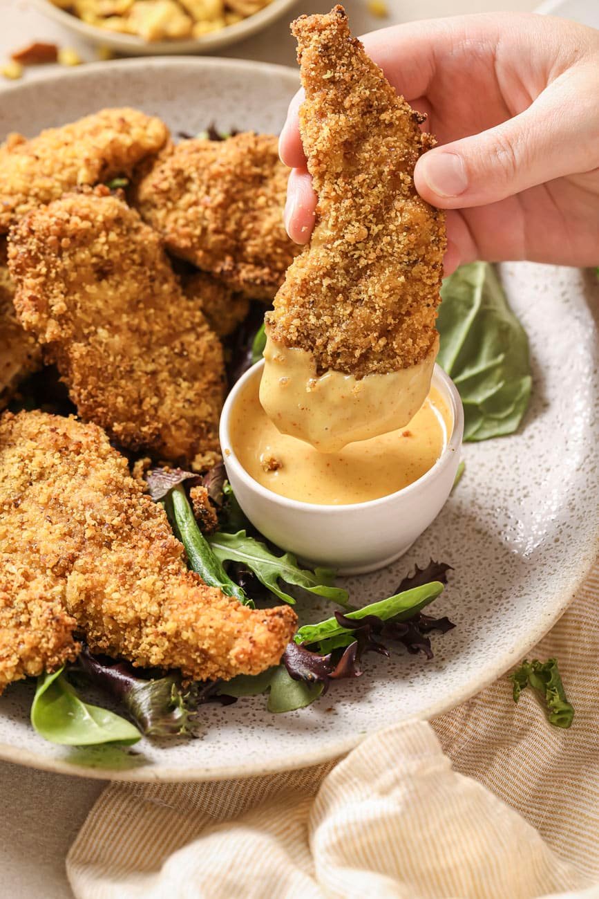 a close up photo of dipping a pretzel crusted chicken tender in honey mustard.