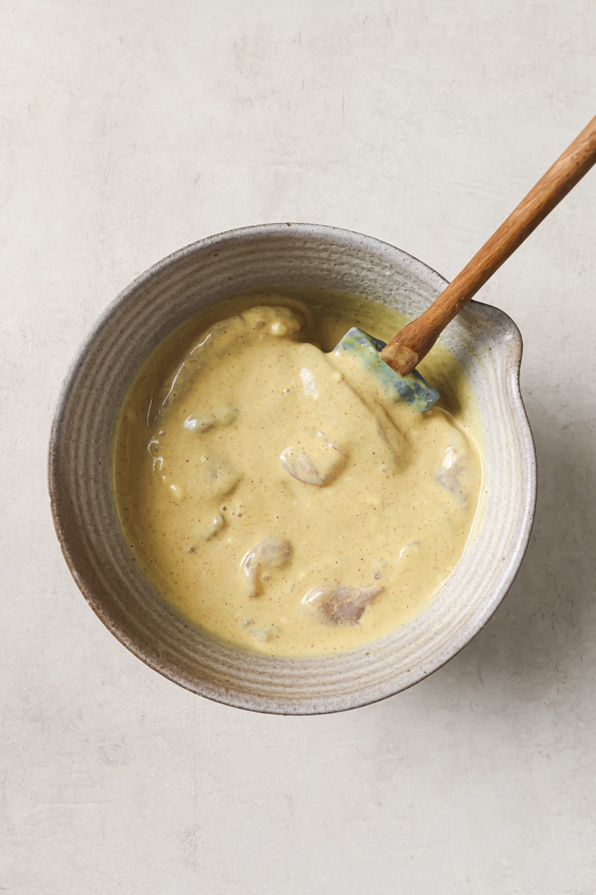 chicken marinating in buttermilk and honey mustard in a mixing bowl with a wooden spatula.