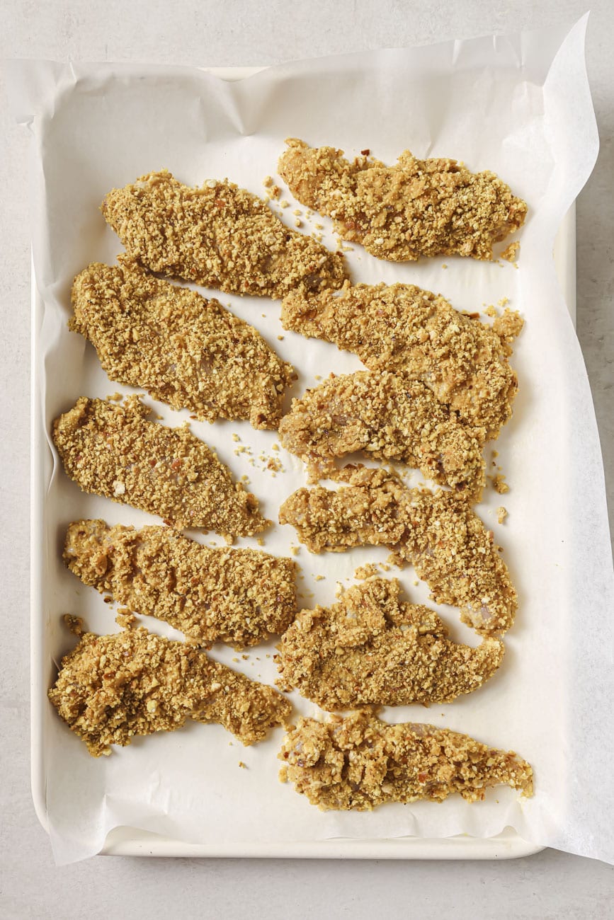 pretzel crusted chicken tenders on a parchment paper lined baking sheet before cooking.