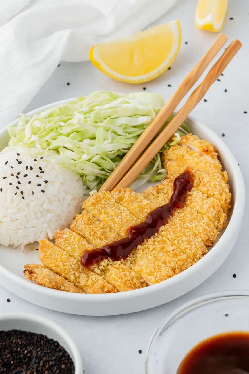 Air fryer chicken katsu on a white plate with rice, chopsticks and lemon slices on the side.