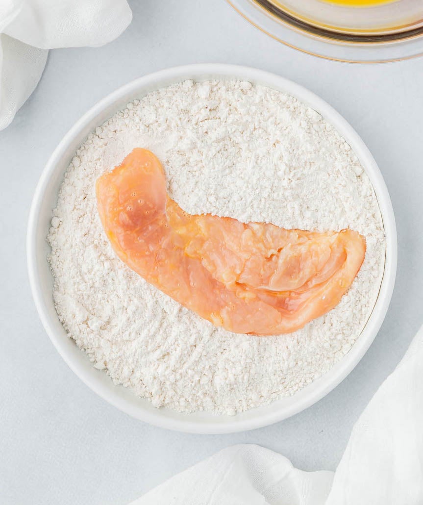 A chicken cutlet in a white bowl filled with flour.